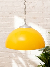Load image into Gallery viewer, A bright yellow dome-shaped pendant light hanging from a silver metal chain against a white brick wall, with a green plant leaf visible in the lower right corner.
