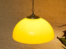 Load image into Gallery viewer, A glowing yellow dome-shaped pendant light hanging against a warm-toned brick wall, with a dark metal top cap and chain visible.