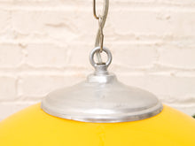 Load image into Gallery viewer, A close-up of a hanging pendant light with a bright yellow globe shade and a brushed silver metal ceiling mount against a white brick wall background.