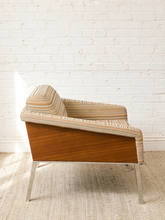 Load image into Gallery viewer, A mid-century modern accent chair with a warm wood veneer side panel and striped fabric upholstery, set against a white brick wall and light beige rug.