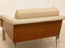 Load image into Gallery viewer, A mid-century modern armchair with a warm brown wood frame and striped fabric upholstery in beige, orange, and blue, shown from a side angle against a white brick wall.