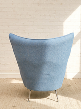 Load image into Gallery viewer, A mid-century modern wingback armchair upholstered in textured light blue fabric, shown from the back on a woven rug against a white brick wall.