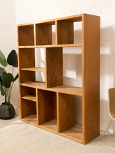 Load image into Gallery viewer, A tall, multi-compartment wooden bookcase with a warm medium-brown finish, styled in a room setting against a white brick wall with a potted plant on the left and a chair on the right.