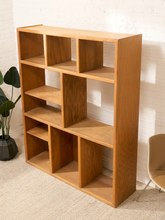 Load image into Gallery viewer, A tall, multi-compartment wooden bookcase with a warm medium-brown oak finish, styled in a room setting with a white brick wall, beige carpet, a potted plant, and a chair.