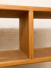 Load image into Gallery viewer, Close-up of a warm medium-brown wooden shelf or cabinet corner with visible wood grain, set against a white painted brick wall.