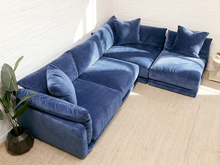Load image into Gallery viewer, A modern sectional sofa upholstered in deep blue velvet fabric, styled in a minimalist room with a white brick wall, beige textured rug, and a potted green plant.
