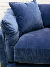Load image into Gallery viewer, A close-up of a blue velvet sofa with matching blue throw pillows, set against a white brick wall background.
