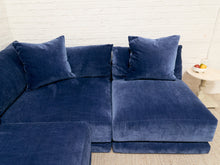 Load image into Gallery viewer, A modern modular sectional sofa upholstered in deep navy blue velvet, featuring matching throw pillows, set against a white brick wall background.