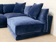 Load image into Gallery viewer, A modern low-profile sectional sofa upholstered in deep navy blue velvet fabric, featuring plush throw pillows and a chaise lounge, set against a white brick wall and beige carpet.