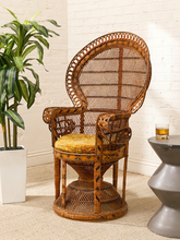 Load image into Gallery viewer, A tall woven rattan accent chair with a high oval backrest, carved details, and a yellow cushion on the seat, set in a room with a white brick wall, a potted plant, and a small side table.