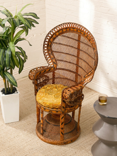 Load image into Gallery viewer, A tall woven rattan accent chair with a high oval backrest and a yellow patterned cushion, styled in a bright room next to a potted plant and a small side table.
