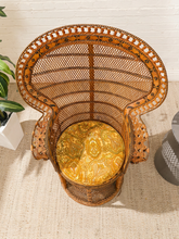 Load image into Gallery viewer, A tall, woven rattan accent chair with a curved backrest and a round cushion featuring a yellow and gold paisley pattern, set on a textured beige rug.