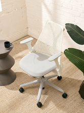 Load image into Gallery viewer, A modern white mesh office chair with a five-star wheeled base, styled in a bright room with a white brick wall, a woven rug, and a potted plant.