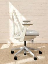 Load image into Gallery viewer, A modern white ergonomic office chair with a mesh back, adjustable armrests, and a five-star wheeled base, positioned in front of a white brick wall on a beige rug with soft sunlight casting shadows.