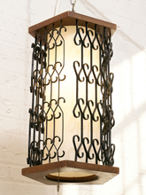 Load image into Gallery viewer, A hanging lantern-style pendant light with a warm white glowing shade, dark metal scrollwork, and medium brown wooden trim, shown against a white painted brick wall.