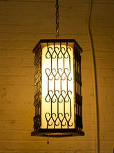Load image into Gallery viewer, A hanging lantern-style pendant light with a glowing warm white frosted center panel and dark metal scrollwork sides, suspended against a yellow-lit brick wall.