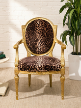 Load image into Gallery viewer, An ornate gold armchair with leopard-print upholstery on the backrest and seat, set against a white brick wall with a potted plant and rug.
