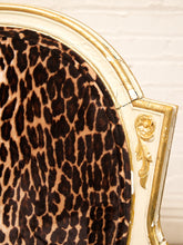 Load image into Gallery viewer, Close-up of an upholstered chair corner showing a leopard-print fabric in tan, brown, and black with a gilded gold frame against a white brick wall.