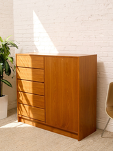 Load image into Gallery viewer, A mid-century modern wooden chest of drawers with five drawers on the left and a single cabinet door on the right, made of warm medium-brown wood, set against a white brick wall with sunlight streaming in.