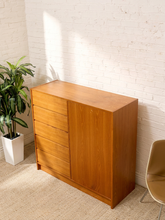 Load image into Gallery viewer, A mid-century modern wooden sideboard with four drawers on the left and a single cabinet door on the right, made of warm medium-brown wood, set against a white brick wall with a potted plant on the left and a chair on the right.