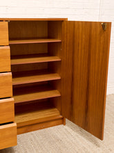 Load image into Gallery viewer, A mid-century modern wooden cabinet with a warm honey-toned wood finish, featuring a partially open door showing five interior shelves and a set of drawers on the left, set against a white brick wall and beige carpet.