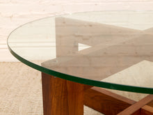 Load image into Gallery viewer, Close-up of a round glass-top table with a wooden base and visible green-tinted glass edge, photographed indoors against a light wall and carpet.