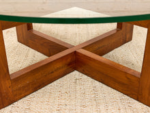 Load image into Gallery viewer, Close-up of a mid-century modern coffee table with a warm brown wooden base featuring an X-shaped cross stretcher and a clear glass top, resting on a woven beige rug.