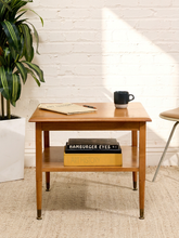 Load image into Gallery viewer, A mid-century modern wooden side table with a warm brown finish, featuring a top shelf and a black-and-white mug on top, set against a white brick wall with a potted plant and a chair in the background.
