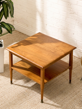 Load image into Gallery viewer, A mid-century modern wooden side table with a warm honey finish, featuring a square top, an open lower shelf, and tapered legs, set on a textured beige rug against a white brick wall.