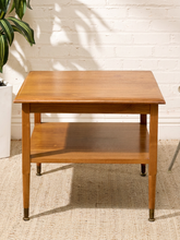 Load image into Gallery viewer, A mid-century modern wooden end table with a warm medium-brown finish, featuring a single shelf and tapered legs with brass feet, set against a white brick wall and a woven jute rug.