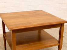 Load image into Gallery viewer, A mid-century modern wooden side table with a warm medium-brown finish, featuring a square top, a lower shelf, and tapered legs, set against a white brick wall.