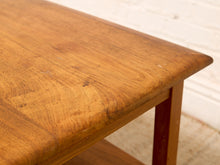 Load image into Gallery viewer, Close-up of a warm medium-brown wooden table corner showing the grain and finish, with part of the leg visible against a white brick wall background.