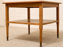 Load image into Gallery viewer, A mid-century modern wooden end table with a warm medium-brown finish, featuring a single shelf and tapered legs with dark metal caps, set on a textured beige carpet against a white brick wall.