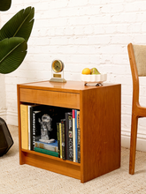 Load image into Gallery viewer, A mid-century modern wooden end table with a warm medium-brown finish, styled with books, a small clock, and fruit against a white brick wall.
