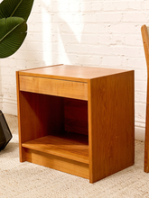 Load image into Gallery viewer, A mid-century modern wooden nightstand with a warm honey finish, featuring a single drawer, an open shelf, and a lower step base, set against a white brick wall.