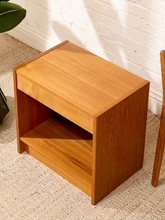 Load image into Gallery viewer, A mid-century modern wooden side table with a warm honey finish, featuring a single drawer and an open shelf, set on a textured beige rug against a white brick wall.