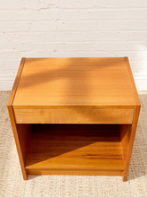 Load image into Gallery viewer, A mid-century modern wooden nightstand with a warm honey finish, featuring a single drawer and an open shelf, set against a white brick wall and light woven rug.