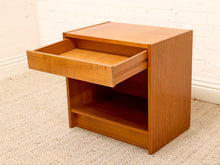 Load image into Gallery viewer, A mid-century modern wooden nightstand with a warm honey finish, featuring an open drawer pulled out and an open shelf below, set on a textured beige carpet against a white brick wall.