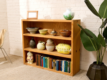 Load image into Gallery viewer, A three-tier wooden bookshelf in a warm oak finish, styled with books, ceramic vases, bowls, and a small picture frame, set against a white brick wall with a potted plant nearby.