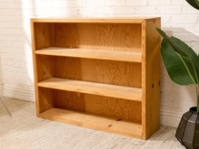 Load image into Gallery viewer, A three-tier wooden bookshelf with a natural light oak finish, positioned against a white brick wall on a beige carpet next to a potted green plant.