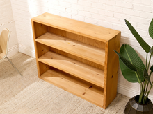 Load image into Gallery viewer, A three-tier wooden bookshelf in a light natural pine finish, styled in a room corner against a white brick wall with a potted plant and a chair partially visible.