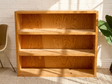 Load image into Gallery viewer, A three-tier wooden bookshelf with a warm honey oak finish, standing against a white brick wall with natural light casting shadows.
