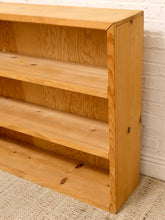 Load image into Gallery viewer, A light natural wood bookshelf with three open shelves, shown in a close-up corner view against a white brick wall and beige carpet.