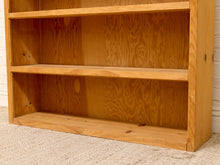 Load image into Gallery viewer, A close-up of a light natural wood bookshelf with three open shelves, standing on a beige carpet against a white brick wall.