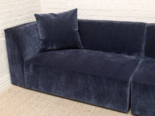 Load image into Gallery viewer, A modern sectional sofa upholstered in dark navy blue velvet fabric, featuring a matching square throw pillow, set against a white brick wall and a light beige woven rug.