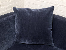 Load image into Gallery viewer, A square navy blue velvet throw pillow resting on a matching navy blue curved sofa in front of a white brick wall.