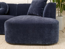 Load image into Gallery viewer, A navy blue velvet modular sectional sofa with a rounded ottoman-style seat and matching back cushions, shown in a styled interior setting with a plant and woven rug.