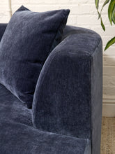 Load image into Gallery viewer, A close-up of a dark navy blue velvet sofa with a matching throw pillow, set against a white brick wall background with a green plant leaf visible in the upper right.