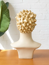 Load image into Gallery viewer, A textured, metallic gold spiky wig displayed on a cream-colored mannequin bust, set on a wooden table against a white brick wall with a green leaf in the background.