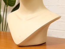 Load image into Gallery viewer, A cream-colored mannequin bust neck display stand on a wooden table with a white brick wall and a green plant in the background.